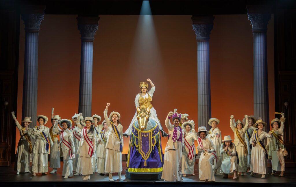 Monica Tulia Ramirez as Inez Milholland stands atop a pedestal, surrounded by the SUFFS Company in costumes.