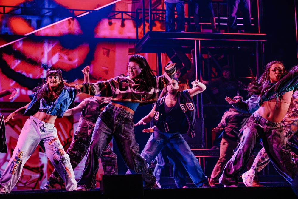 A group of dancers performs energetically on stage, wearing colorful outfits against a vibrant backdrop.