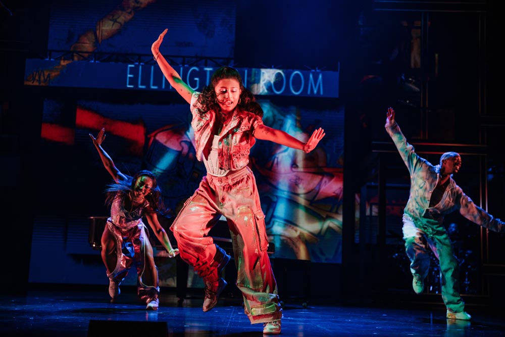 Dancers perform energetically on stage, wearing colorful costumes, with vibrant lighting and abstract projections in the background.