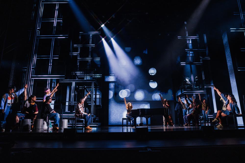 A group of performers on stage, with a piano, under dramatic lighting and a modern backdrop.