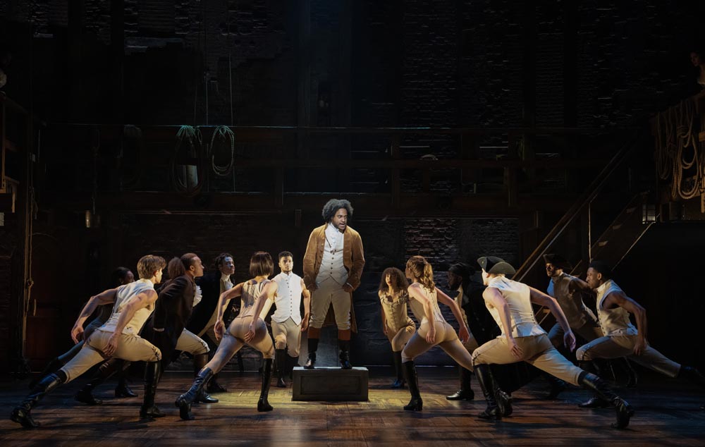 Tyler Fauntleroy stands center stage, surrounded by dancers in period costumes, under dramatic lighting.