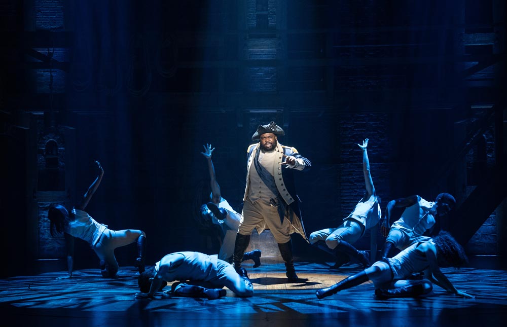 A.D. Weaver performs center stage in a dramatic pose, surrounded by dancers in white costumes under blue lighting.