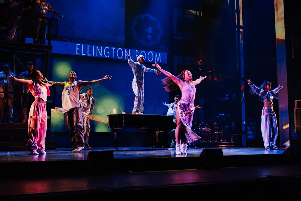 The cast performs energetically on stage, with vibrant costumes and a grand piano in a colorful setting.