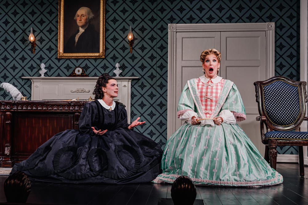 Betty Gilpin and Bianca Leigh perform in period dresses, expressing emotion against a patterned wallpaper backdrop.