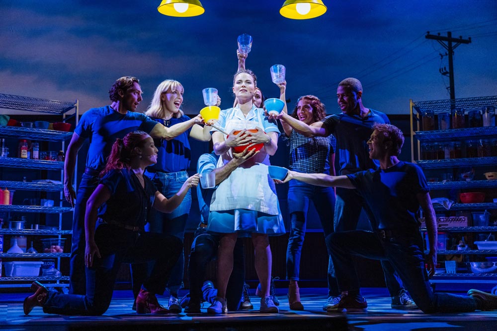 A waitress in a white dress holds a pie, surrounded by six performers in dark attire.