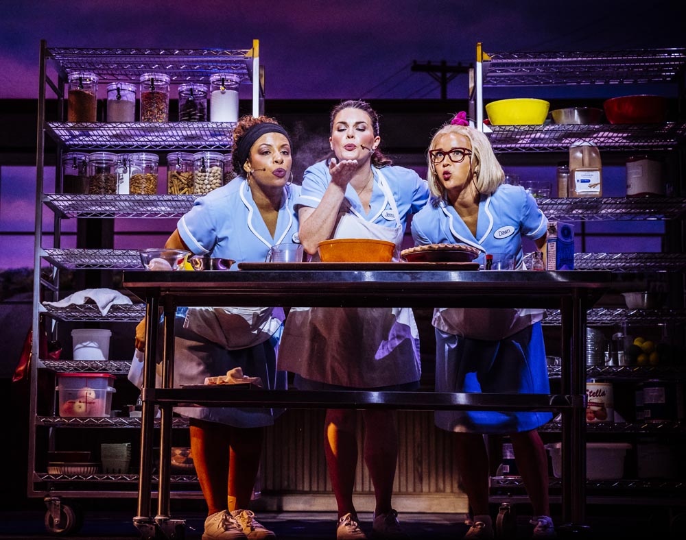Three waitresses in blue uniforms lean over a table, playfully interacting with a pie, surrounded by kitchen shelves.