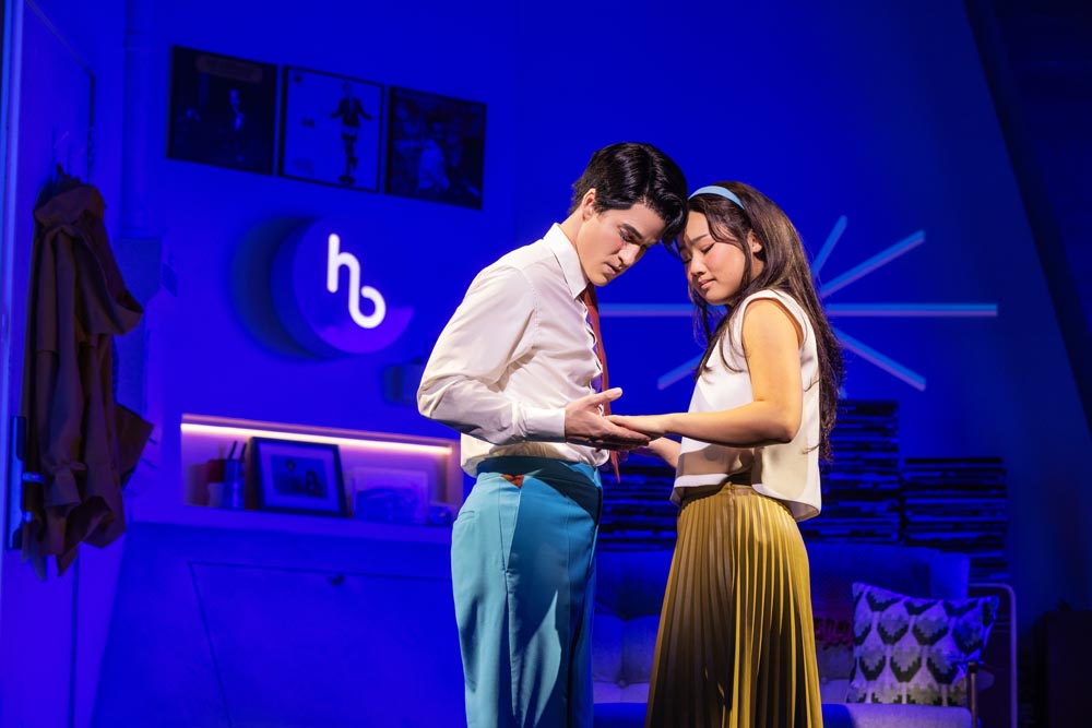 Darren Criss and Helen J. Shen share an intimate moment on stage, illuminated by colorful lighting.