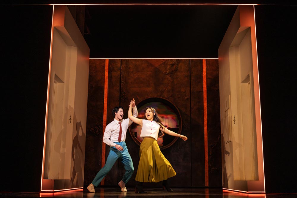 Darren Criss and Helen J. Shen dance joyfully on stage, surrounded by a vibrant, theatrical backdrop.