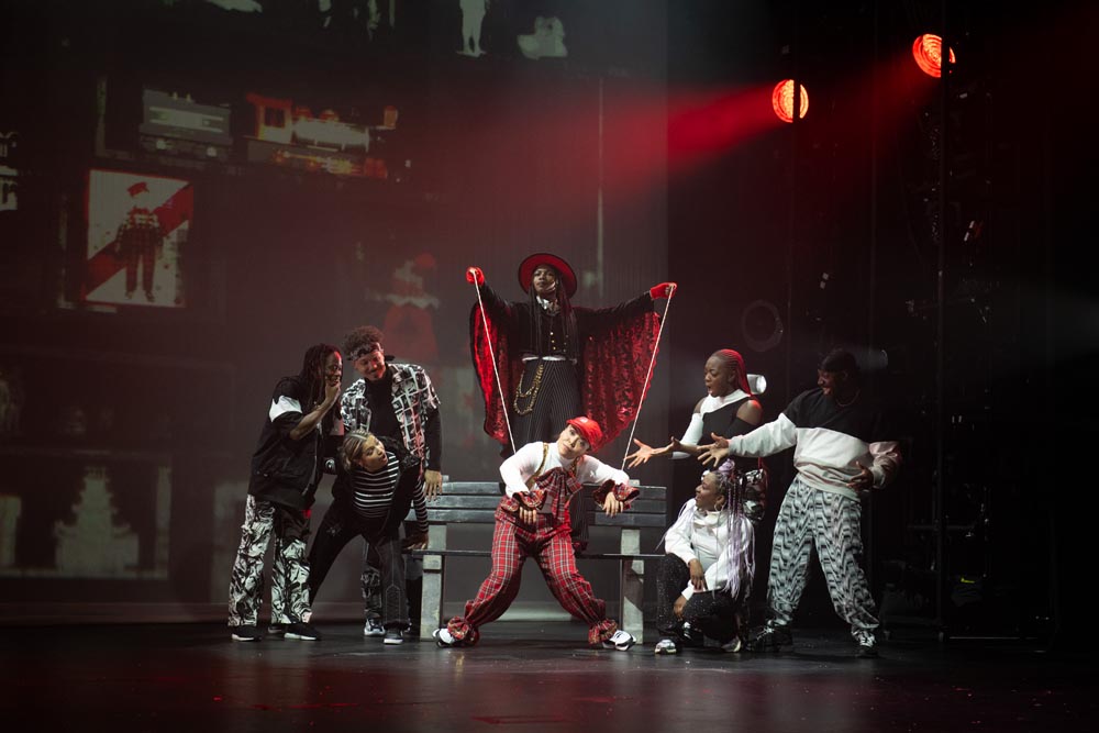 A performer in a red cape poses dramatically, surrounded by dancers in black and white outfits.