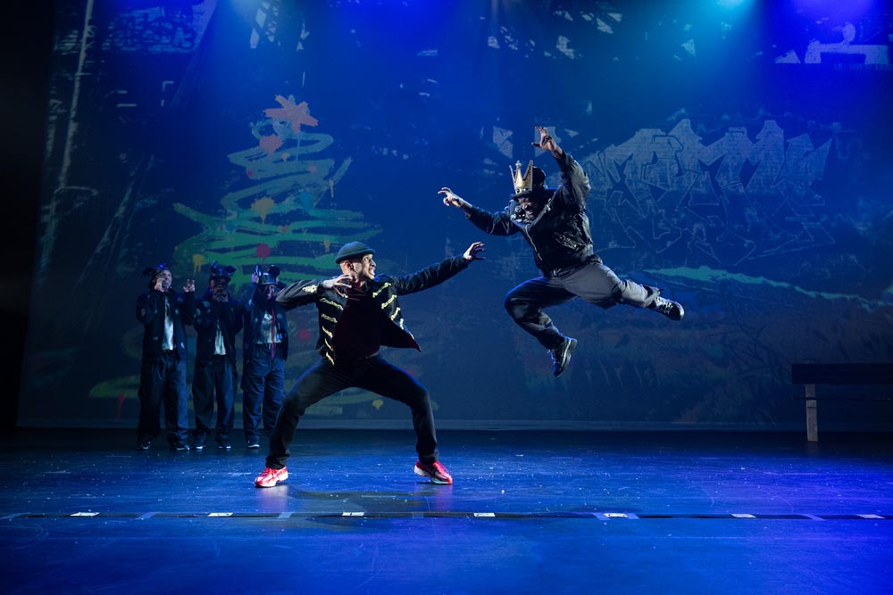 Two dancers perform a dynamic hip-hop move on stage, with colorful lights and a cheering audience in the background.