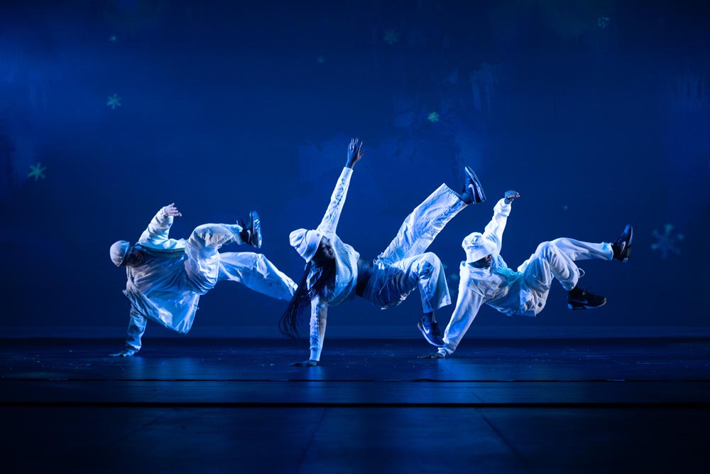Four dancers perform dynamic moves on stage, wearing white outfits against a blue-lit background.