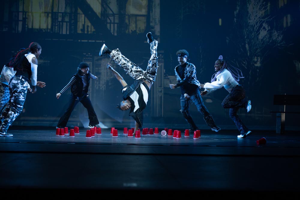 A dancer performs a handstand surrounded by three others, with red cups scattered on the stage.