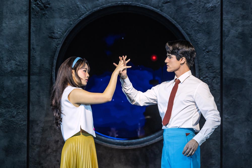 Helen J. Shen and Darren Criss touch hands through a circular backdrop, wearing colorful costumes.