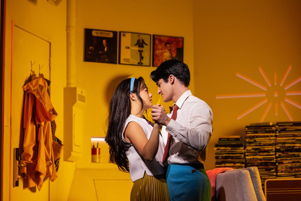 Helen J. Shen and Darren Criss dance closely, illuminated by warm yellow light, with records in the background.