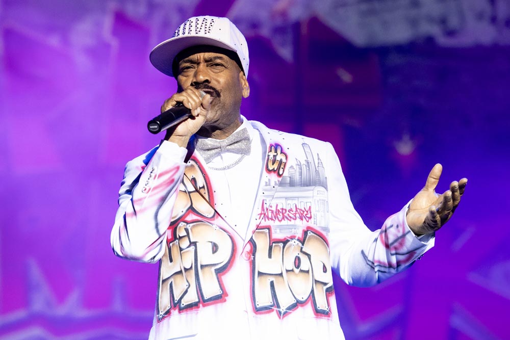 A performer in a white jacket with colorful graffiti-style "HIP HOP" sings into a microphone.