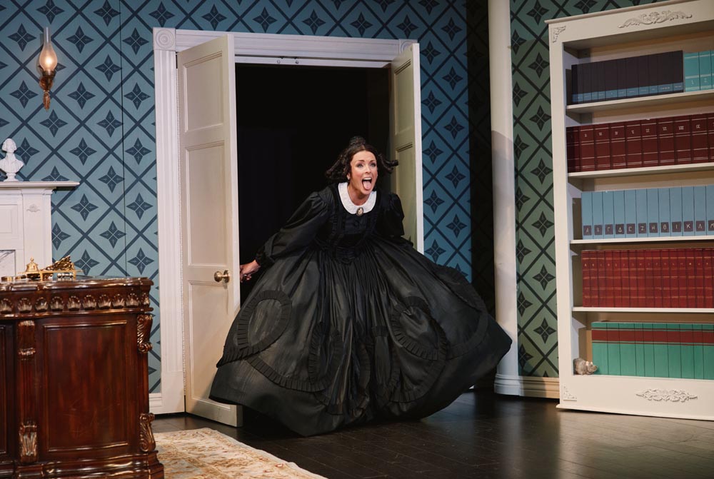 Jane Krakowski in a black dress, joyfully entering through a doorway, with a patterned blue background.