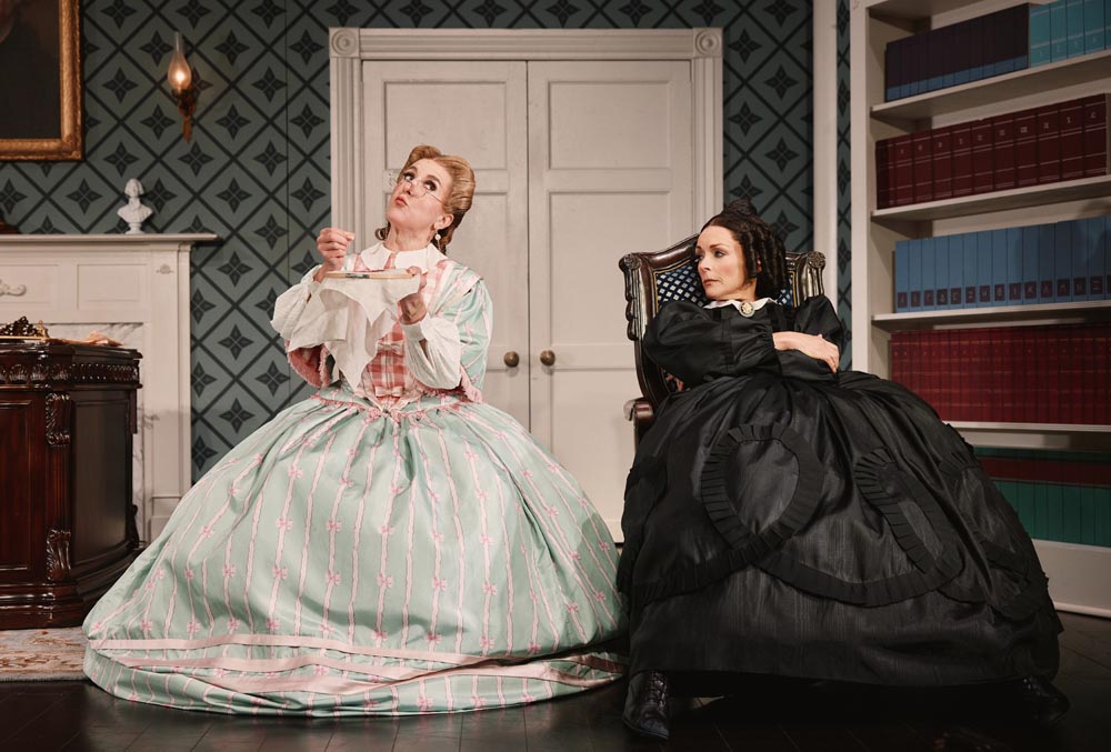 Jenn Harris and Jane Krakowski in period costumes, seated in a room with patterned wallpaper.