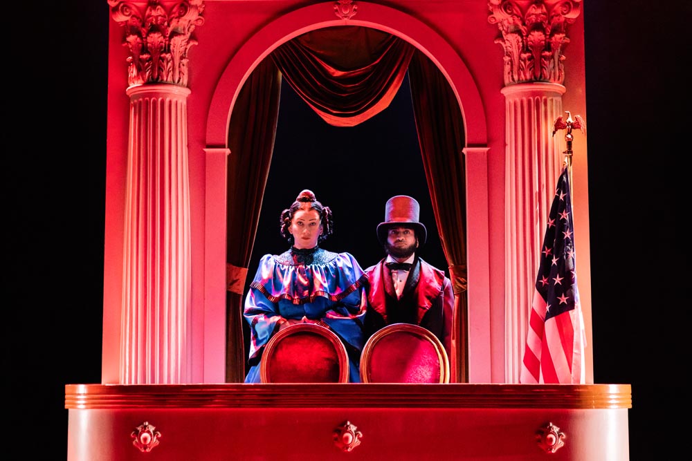 Jinkx Monsoon and Kumail Nanjiani seated in a theater box, surrounded by red curtains and decor.