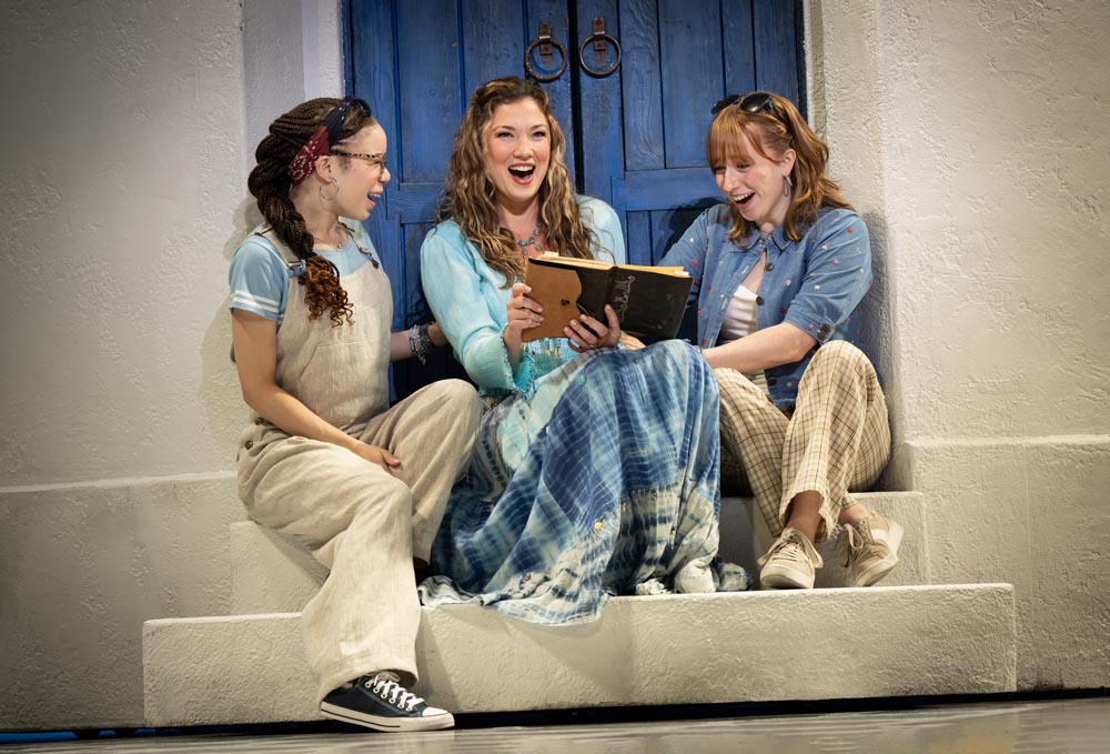 Three women sit together, laughing and sharing a book, with a blue door in the background.