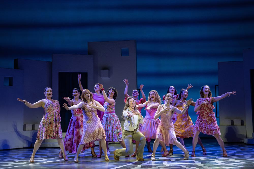 A vibrant ensemble cast in colorful dresses performs energetically on stage, with a blue backdrop.