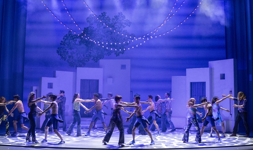 A group of dancers in casual attire perform on stage, surrounded by blue lighting and decorative strings.