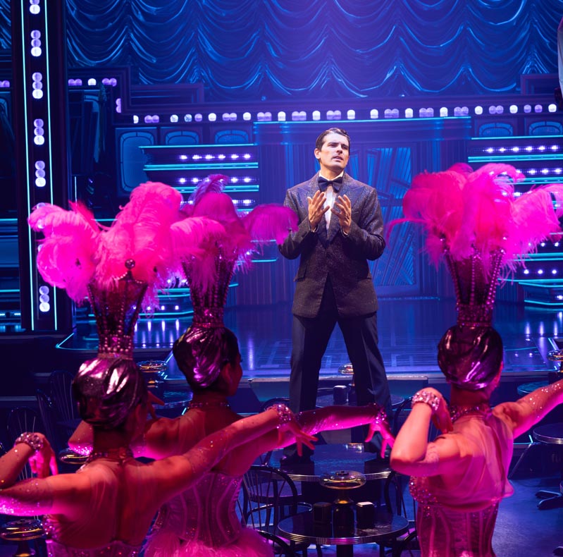 Matt Magnusson as Bobby Darin performs on stage, surrounded by dancers in pink feathered headdresses.
