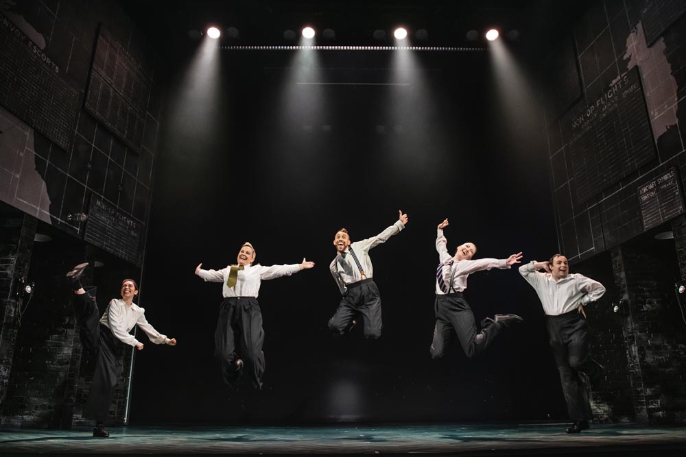Five performers in white shirts and black trousers leap joyfully on stage under bright spotlighting.