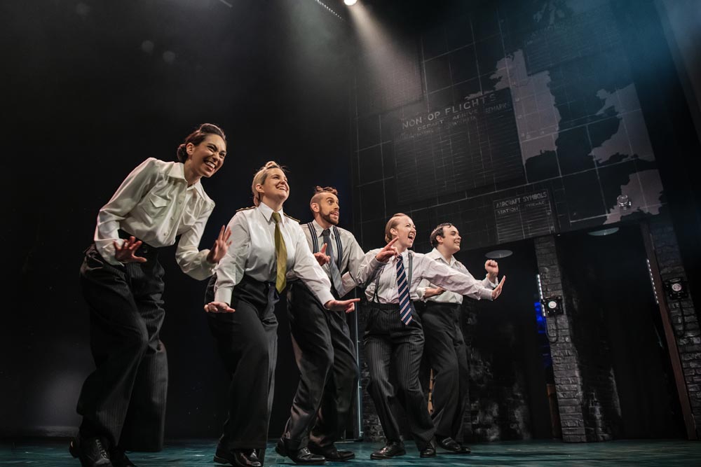 Five performers in formal attire dance energetically on stage, illuminated by dramatic lighting in a theater.