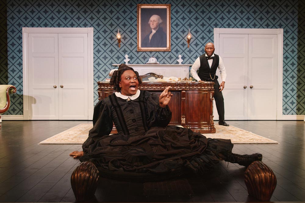 Tituss Burgess, in a black dress, sits on the floor, while Phillip James Brannon stands behind him.